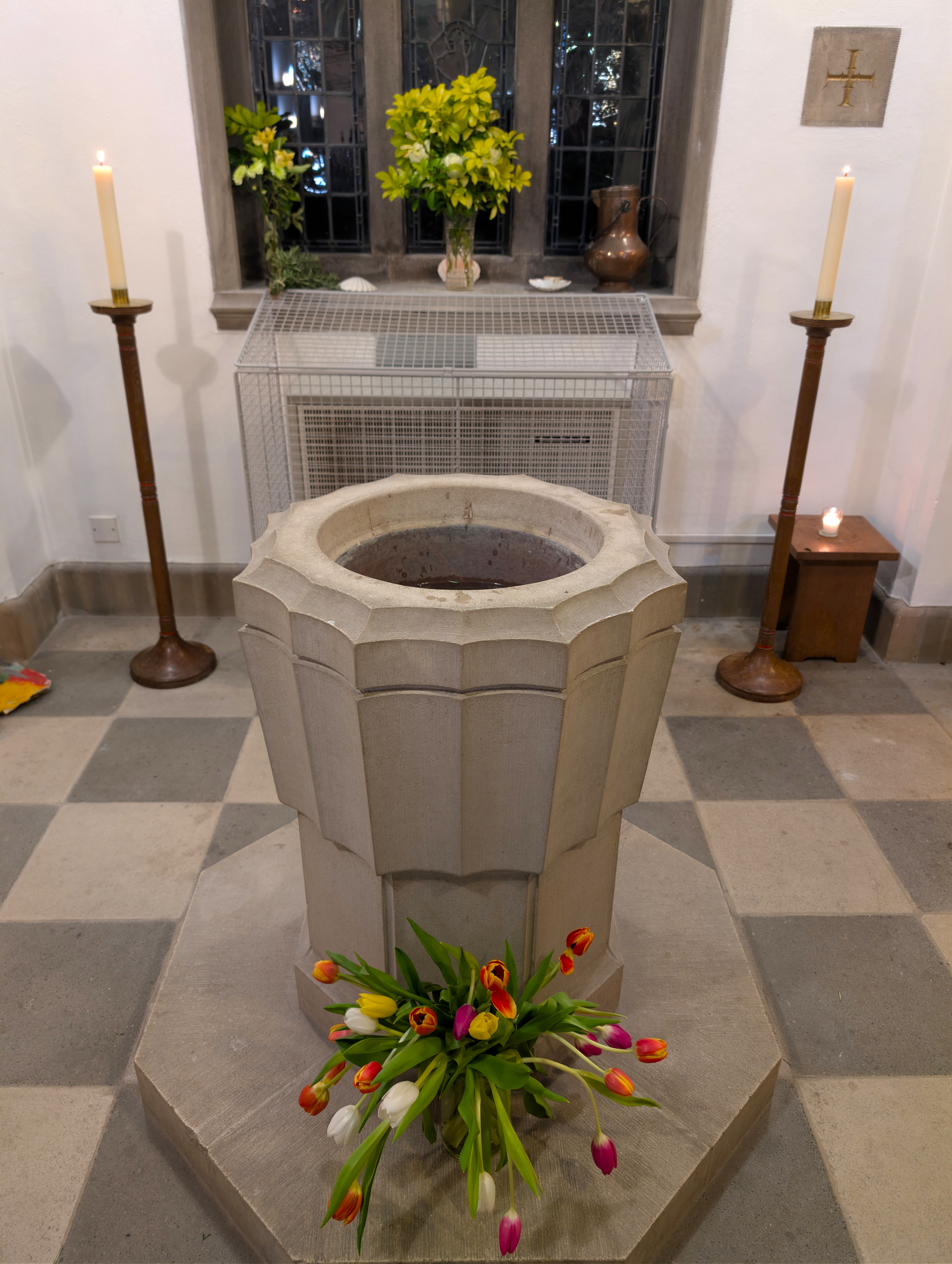The baptismal font within a church, filled with water and surrounded by flowers